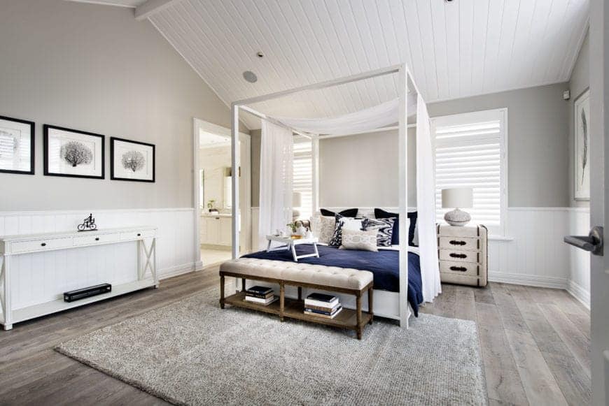 The white wooden four-poster bed pairs well with the white wainscoting and the white wooden finish of the cathedral ceiling that is complemented by the light gray walls and the bright natural lights coming in from the shuttered windows.