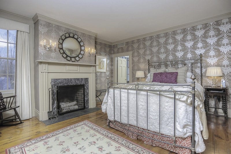 The metal railings of the mission-style bed blend with the subtle silvery patterns of the wallpaper that dominated the walls. This makes the fireplace stand out with its wooden mantle that is adorned with a decorative mirror mounted on the wall above.