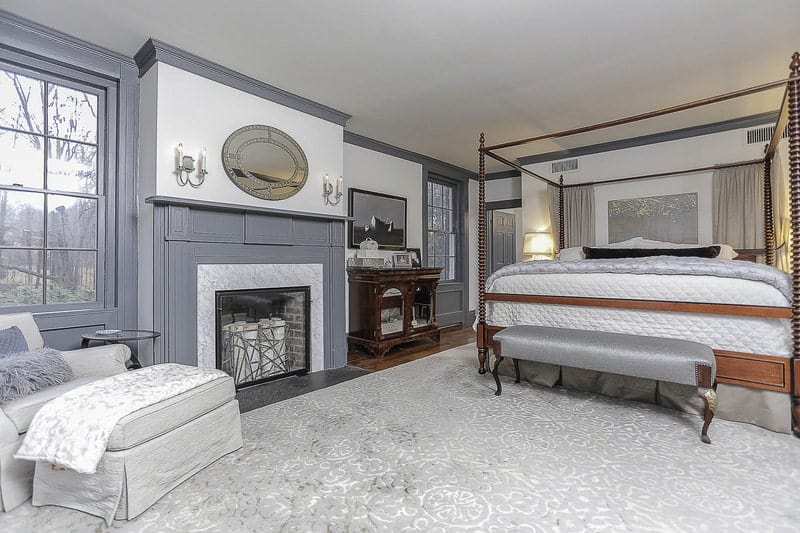 The large wooden four-poster bed dominates this bedroom. It has gray sheets that match the gray finish of the wooden walls and the mantle of the fireplace beside the sitting area that has a light gray cushioned armchair and a foot stool by the window.