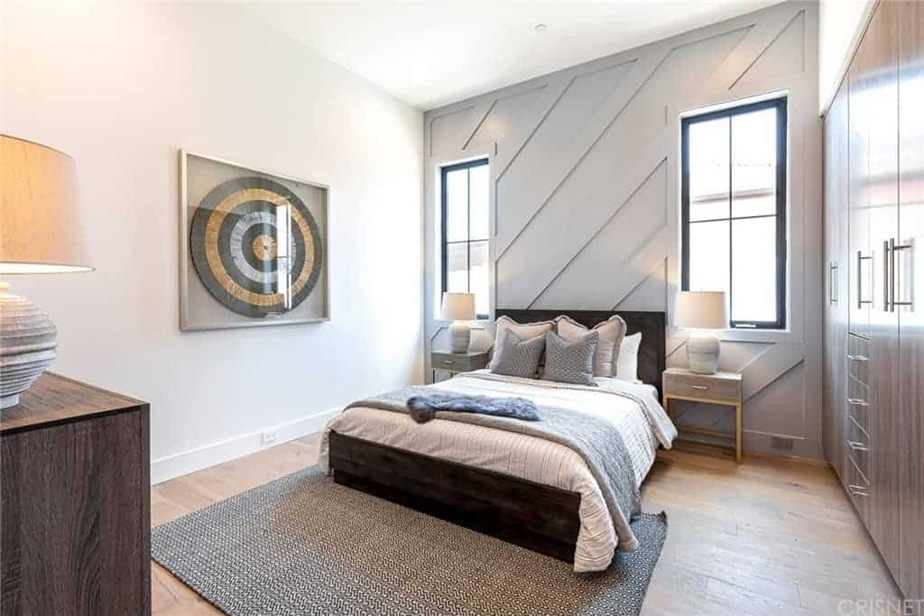 The far wall has a light gray hue and a Farmhouse-style wooden finish to it that pairs well with the dark wooden bed and the hardwood flooring. This bed has a headboard flanked by table lamps on bedside drawers as well as brilliant windows that brighten the white ceiling.