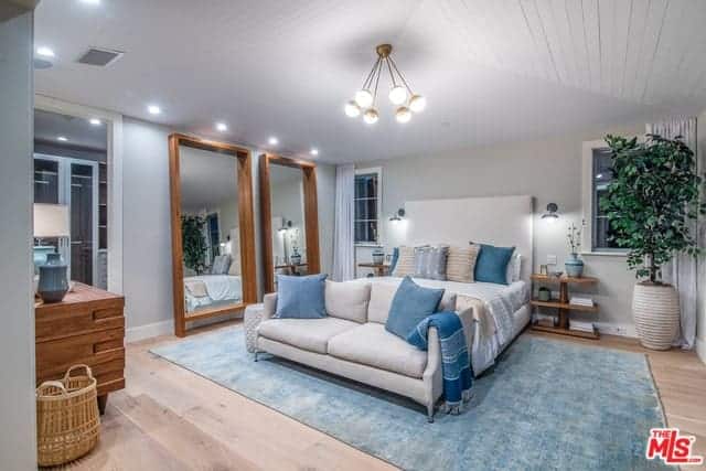 There is a pair of large wood-framed mirrors leaning on the light gray wall beside the bed that has a large cushioned headboard flanked by wall-mounted lamps over wooden bedside tables with a unique design blending with the hardwood flooring that is topped with a light green area rug.