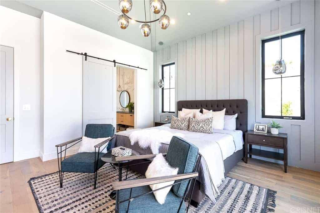 The chocolate brown tufted cushioned headboard of the traditional bed stands out against the white wooden plank finish of the wall behind it. This pairs well with the flanking bedside drawers and the hardwood flooring.