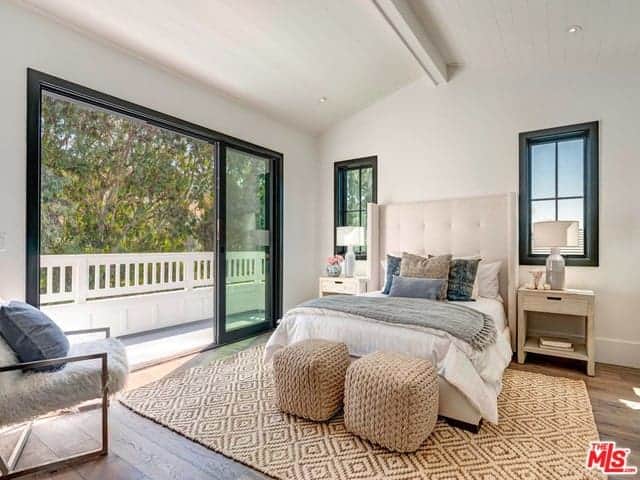 The highlight of this Farmhouse-style bedroom is the rustic woven patterned are rug that covers the hardwood flooring. It matches with the couple of ottomans at the foot of the bed with white sheets and a light beige cushioned headboard.