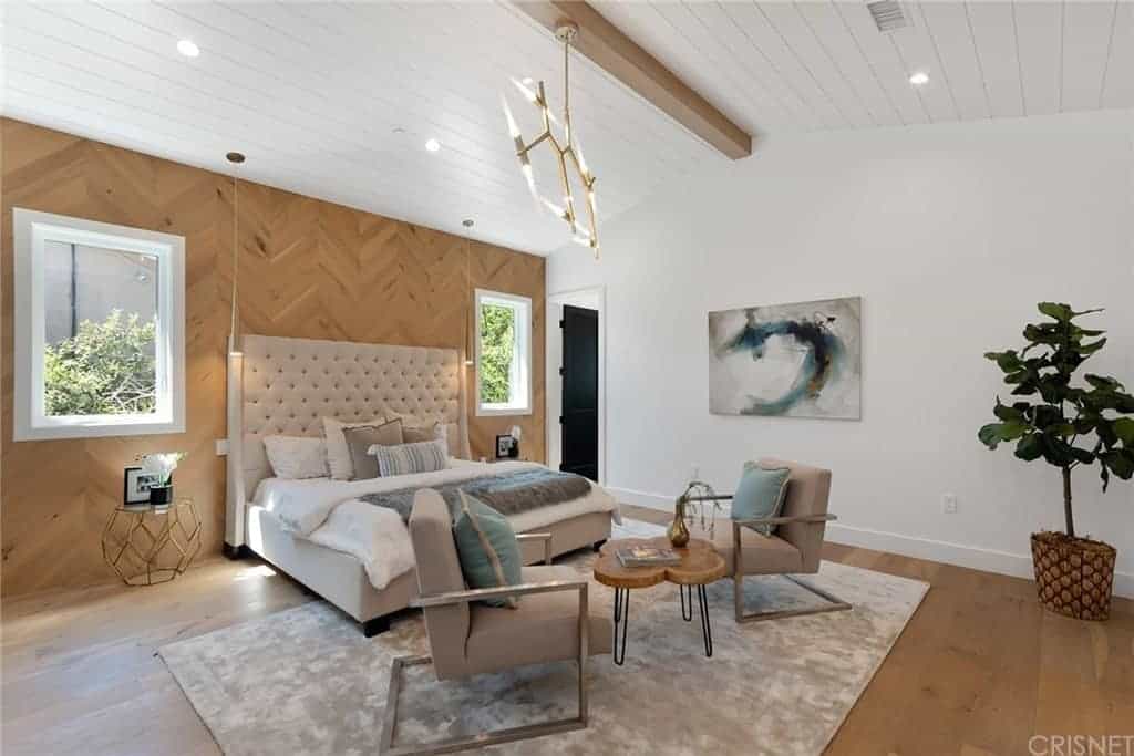 This is a Farmhouse-style bedroom through and through. It has a hardwood flooring that blends with the wooden wall behind the large tufted cushioned headboard. It also matches with the exposed wooden beam running the middle of the white cathedral ceiling that has a wooden plank finish.