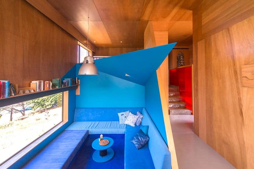 A tiny living room with blue walls and furniture achieving a cohesive look. It is illuminated by a chrome dome pendant that hung from the wood-paneled ceiling.