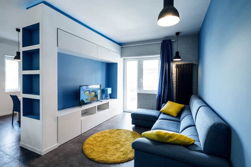 Modern living room boasts a built-in cabinet and a sectional sofa accented with yellow pillows and a round rug. It is lighted by black dome pendants along with natural light from the glazed windows.
