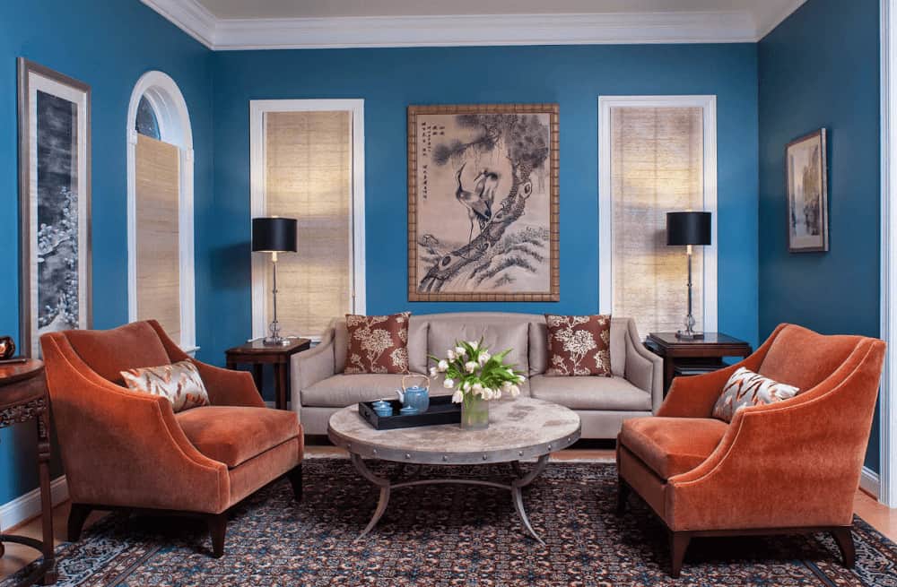 Traditional living room decorated with lovely artworks mounted on the blue walls. It has orange velvet chairs and a taupe sectional paired with a round coffee table that sits on a patterned rug.