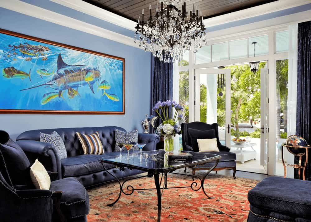 A large fish painting hangs over the leather tufted sofa facing the glass top coffee table over a red area rug. It is illuminated by a glam candle chandelier that hung from the tray ceiling clad in wood planks.