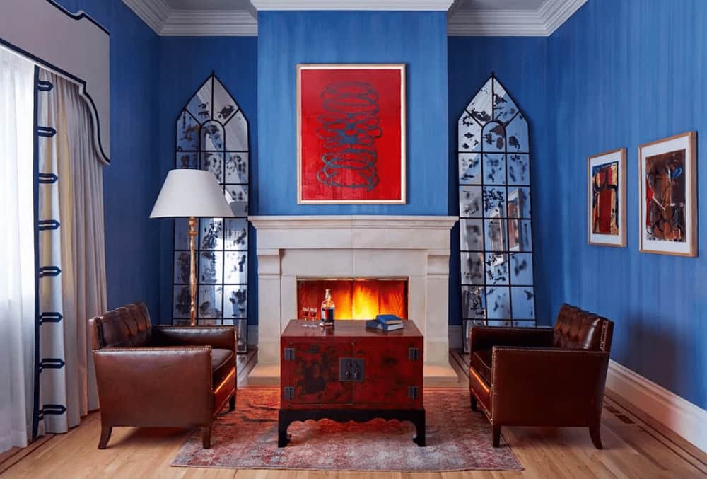 A red artwork stands out against the blue walls lined with white base and crown moldings. It has leather tufted armchairs and a wooden coffee table facing the fireplace flanked by stylish mirrors.