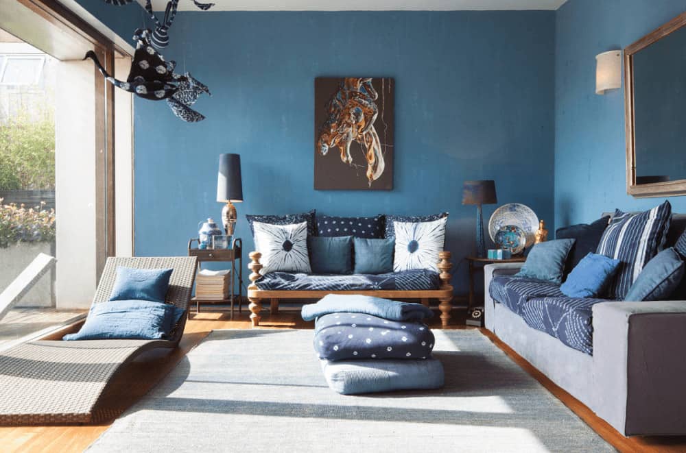 Serene living room furnished with cozy seats and a wicker lounge chair by the full height glazing inviting natural light in. It has interesting artwork and layered floor pillows that sit on a gray area rug.