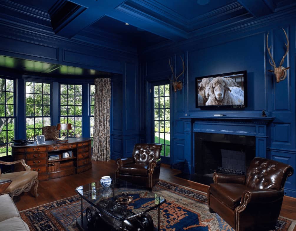 Airy living room with coffered ceiling and full height wainscoting mounted with stag head decors. It has cozy seats and an oval desk that blends in with the rich hardwood flooring.