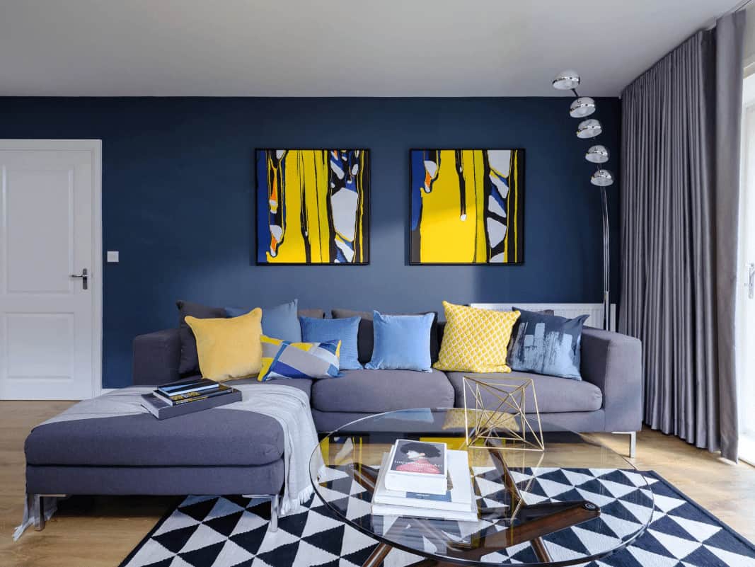 Blue living room decorated with yellow artworks mounted above the L-shaped sofa filled with fluffy pillows. It has a five-head floor lamp and a glass top coffee table that sits on a striking area rug over the light hardwood flooring.
