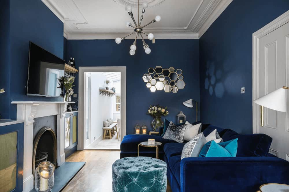 Blue living room decorated with cluster mirror and a contemporary chandelier that hung over the round tufted ottoman. It has an L-shaped sofa and an arched fireplace with a wall mount TV on top.