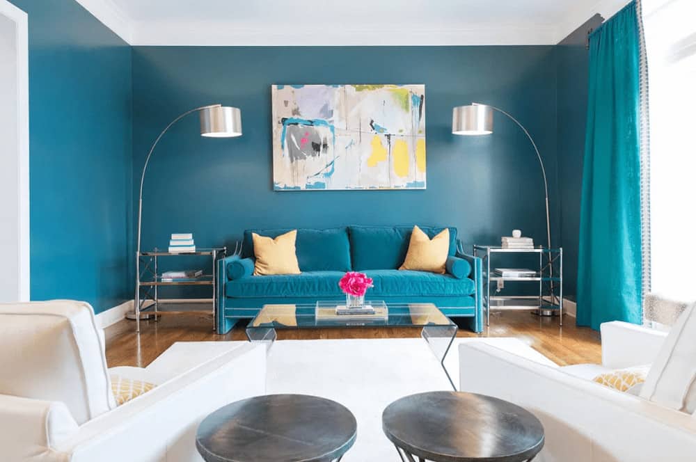 An abstract painting hangs over the blue sofa that blends in with the walls and drapes. It is flanked by metal side tables and chrome floor lamps with drum shades.