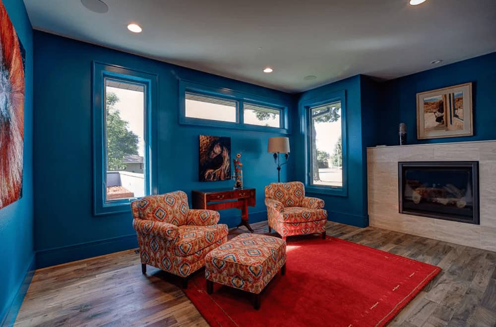 This living room showcases a fireplace and patterned armchairs paired with a matching ottoman over a red area rug. It has natural hardwood flooring and blue walls mounted with artworks.