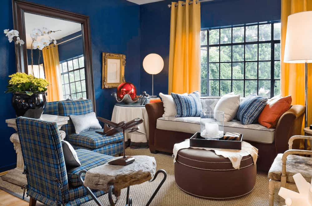 Yellow draperies covering the aluminum-framed window stand out in this blue living room with a large mirror and mismatched seats surrounding a round ottoman that sits on a jute area rug.