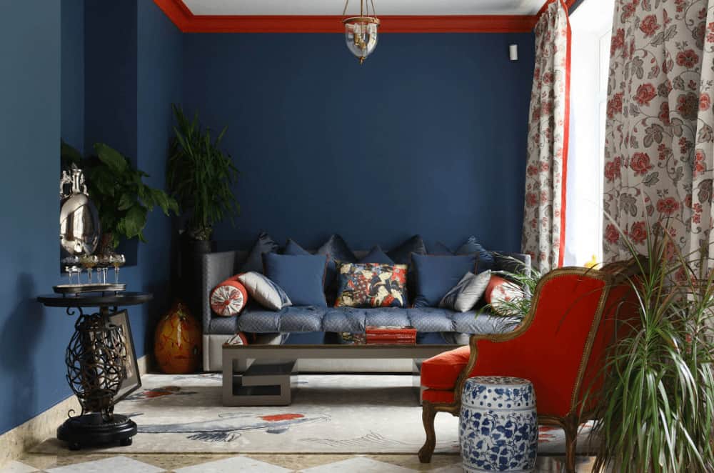 Red crown molding provides a sleek contrast to the blue walls within this living room. It complements the wingback chair accompanied by a cushioned sofa and sleek coffee table.
