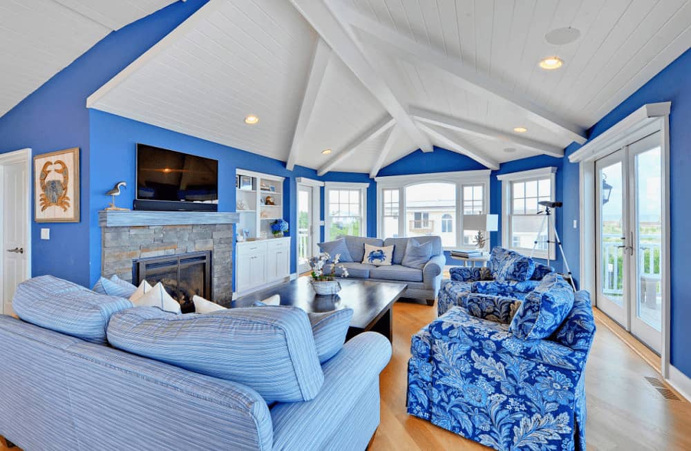 Beach style living room with rich hardwood flooring and shiplap ceiling lined with white beams. It includes a brick fireplace and gorgeous blue seats surrounding a dark wood coffee table.