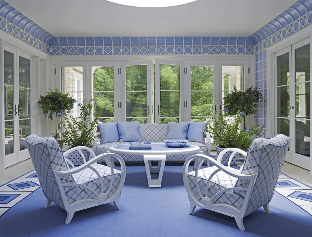 This living room is furnished with a round coffee table and patterned seats that harmoniously blend with the flooring and stylish ceiling. It has potted plants and French doors that open to the lush green yard.