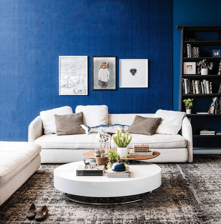 Interesting wall arts hang above the beige sofa paired with a round coffee table over a distressed area rug. It includes black shelving fitted against the blue wall.