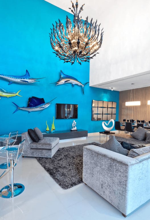 An open living area designed with fish wall decors and a stunning chandelier that hung over the gray shaggy rug. It has velvet seats and a flat-screen TV hanging above the sleek console topped with contemporary sculptures.