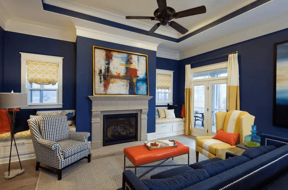 Blue living room with subtle yellow and orange accents from the drapes and valances along with wingback armchair and cushioned coffee table. It has a sectional sofa and a fireplace with gorgeous artwork on top.