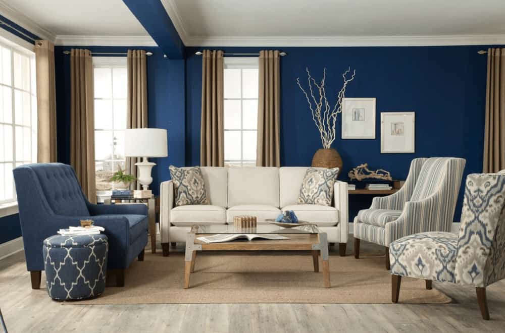 A glass top coffee table sits in the middle of mismatched chairs and beige sectional lighted by a drum table lamp. This room has natural hardwood flooring and white framed windows covered in taupe draperies.