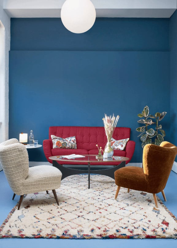 This living room is furnished with mismatched seats and a glass top coffee table illuminated by a round pendant light. It has blue walls and flooring topped by a charming area rug.