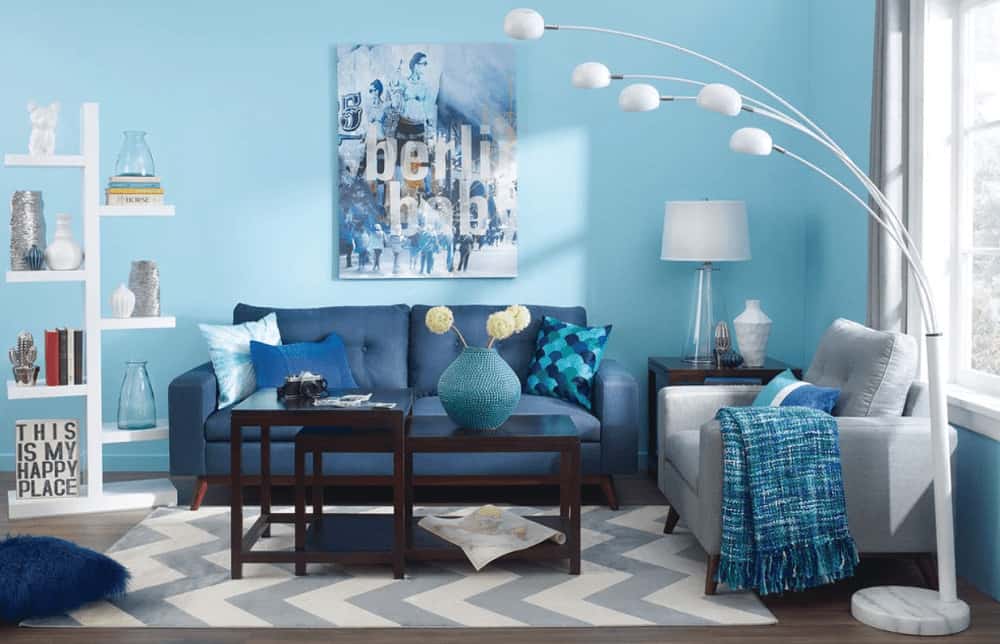 A white shelving unit complements the arched five head floor lamp illuminating the tufted seats and modular coffee table that sits on a gray chevron rug. This room is decorated with lovely artwork mounted on the sky blue wall.