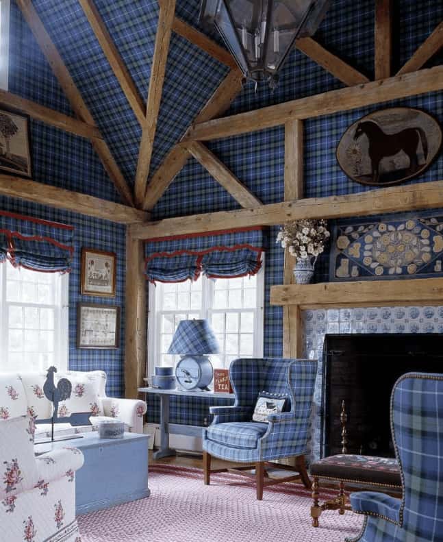 Clad in plaid wallpaper that matches the wingback chairs, this living room offers floral sofas and a fireplace framed in decorative tile surround. It has exposed wood beams adding a rustic touch to the charming room.