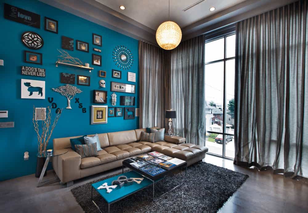 This living room is designed with an eclectic art gallery mounted above the L-shaped sofa that's lighted by a spherical pendant. It has gray shaggy rug and modular coffee table facing the full height glazing covered in sheer curtains.