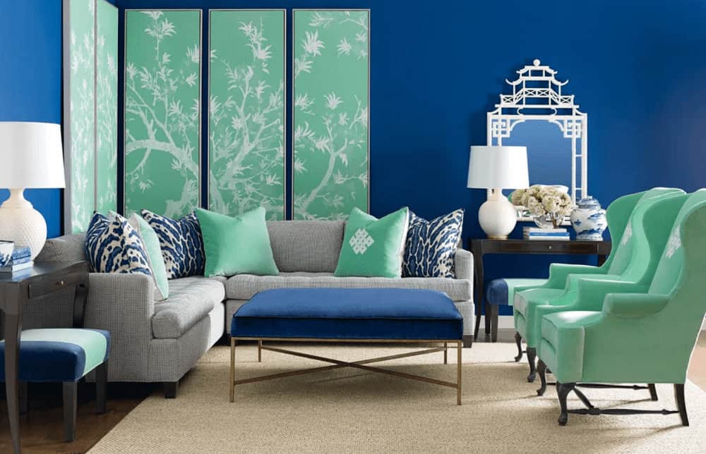 Blue living room with pops of mint greens from the multi-panel wall art, wingback chair and pillows laying on the gray sofa. It has a cushioned coffee table and dark wood side tables topped with white lamps.