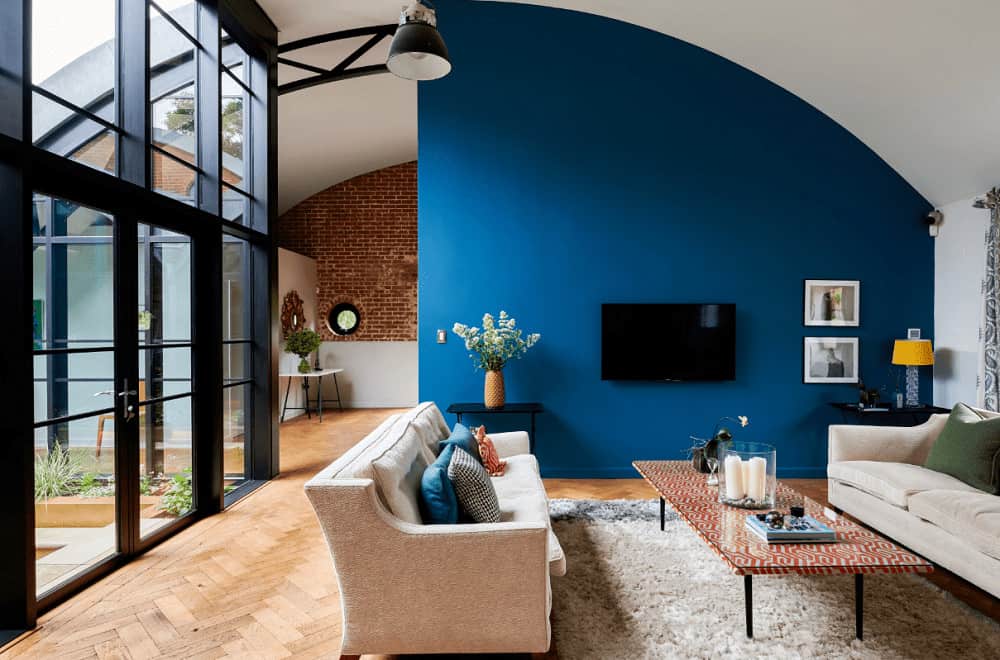 Contemporary living room with arched ceiling and herringbone wood flooring topped by a shaggy rug. It includes beige sectional sofas with a patterned coffee table facing the wall mount TV.