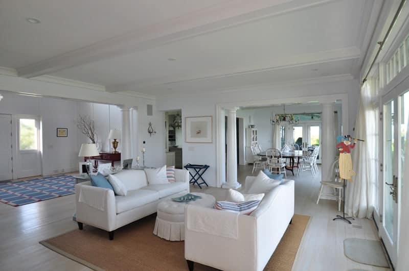 An open living room with a skirted ottoman and facing sofas blending in with the white walls and ceiling. It includes a brown area rug that lays on the wide plank flooring.
