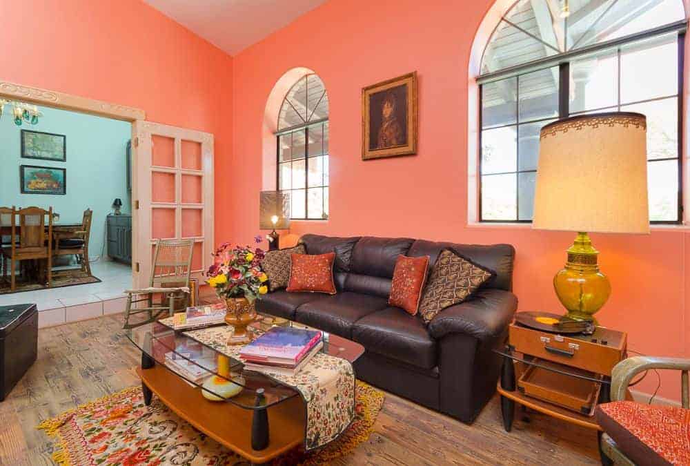 The pink living room showcases a glass top coffee table and a black leather sofa illuminated by oversized table lamps. It has arched windows and hardwood flooring topped by a tasseled rug.