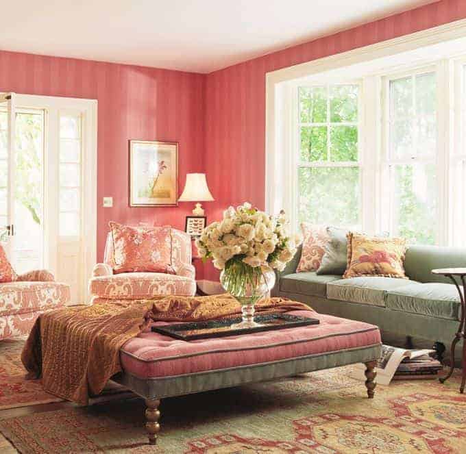 Clad in pink striped wallpaper, this living room features mismatched seats and a tufted ottoman topped with a throw blanket and large flower vase.