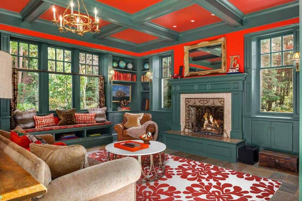 A stylish coffee table sits on a floral area rug complementing with the red pillows and cushion fitted on the window seat nook. This room has a fireplace and brown sofa lighted by a brass chandelier that hung from the coffered ceiling.