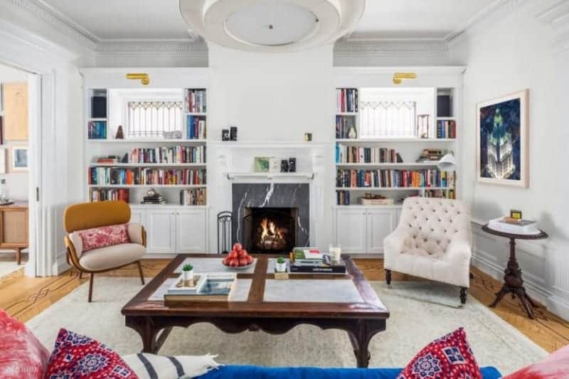 A cozy sofa faces the wooden coffee table and fireplace accompanied by mismatched armchairs. It is flanked by built-in shelvings filled with books.