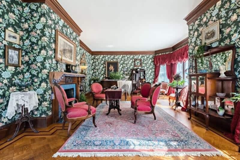 Victorian living room clad in green floral wallpaper that's filled with framed photos. It has a fireplace and red armchairs surrounding a wooden coffee table over a tasseled area rug.