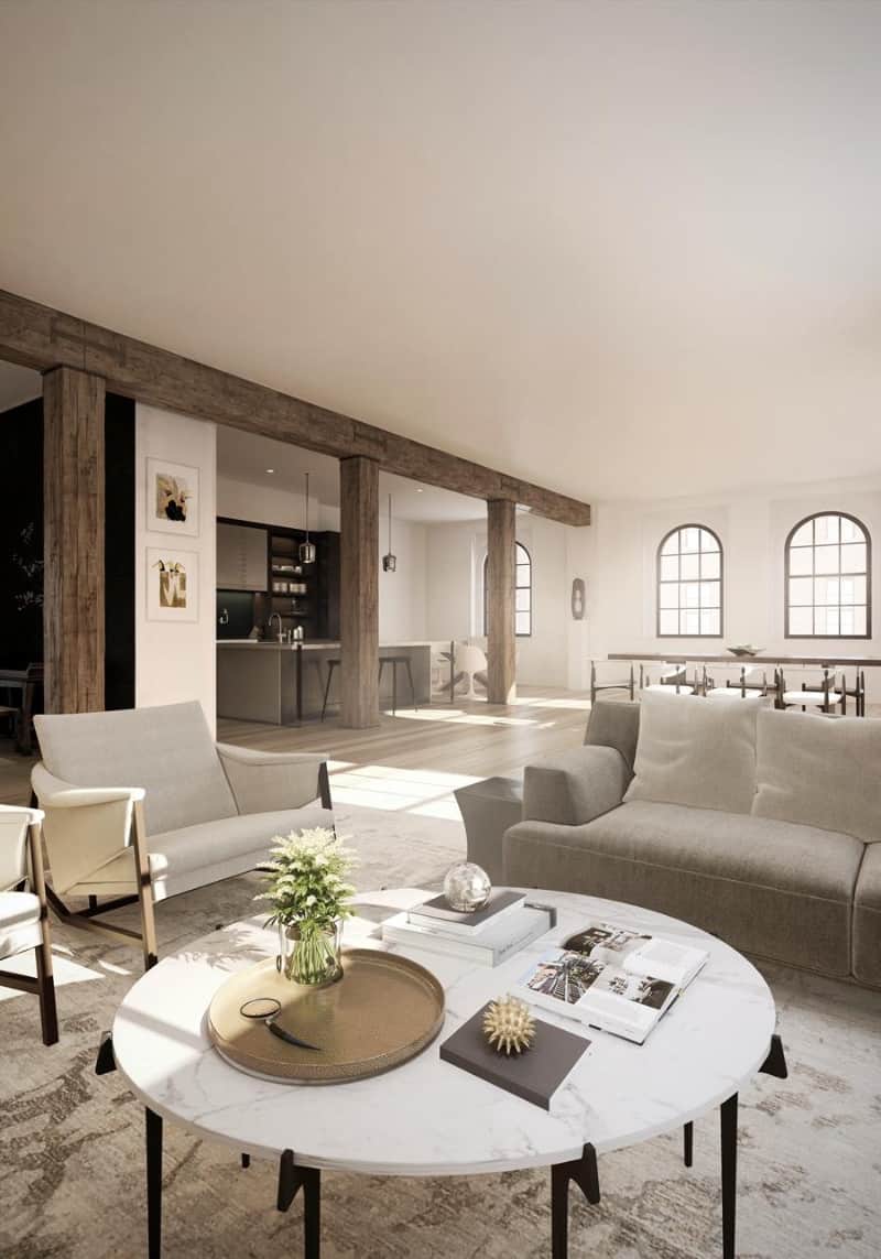 An expansive living room lined with wooden beam and columns offers beige armchairs and a gray sofa paired with a round marble coffee table over a printed area rug.