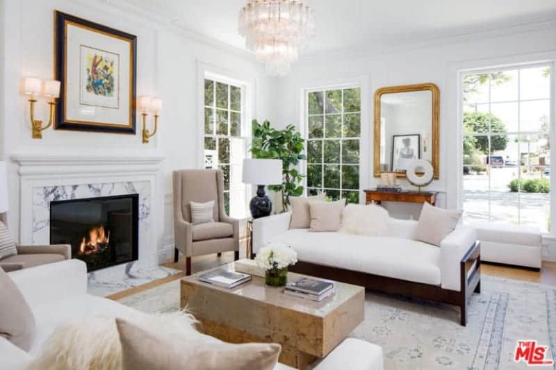 A fancy chandelier illuminates the sleek coffee table situated in between the white sofas filled with neutral and white faux fur pillows. It is accompanied by beige chairs and a fireplace framed in marble surround tiles and white mantel.