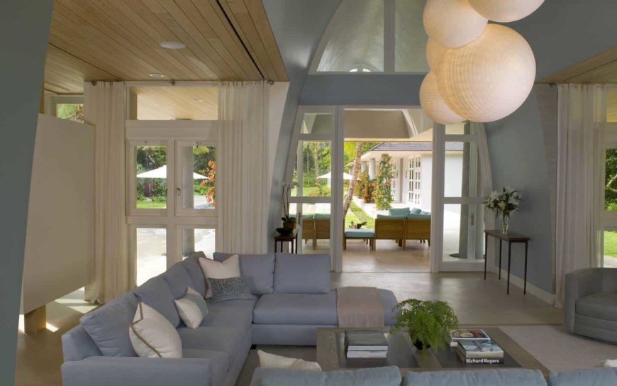 Oversized round pendants hang over the wooden coffee table accompanied by blue sectional sofas and gray armchair. This room has a vaulted ceiling and glass slider that opens to the porch.