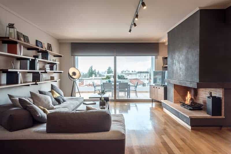 An L-shaped sofa is situated under the floating shelves filled with books and decors. It faces the open fireplace with a flat-screen TV on the side sitting on a wooden console table.