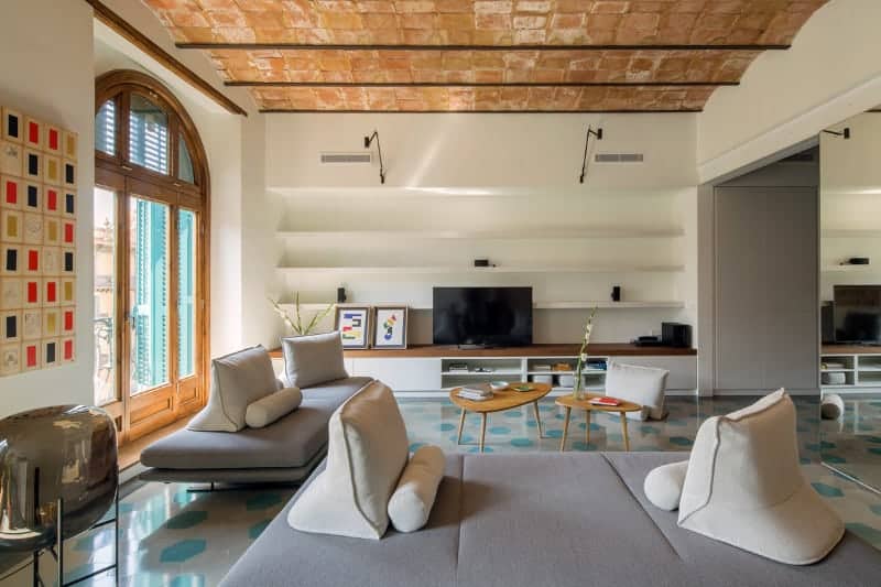 The sleek living room showcases an arched window and barrel vaulted ceiling clad in bricks. It has modern seats and wooden coffee tables facing the TV against the open shelving.