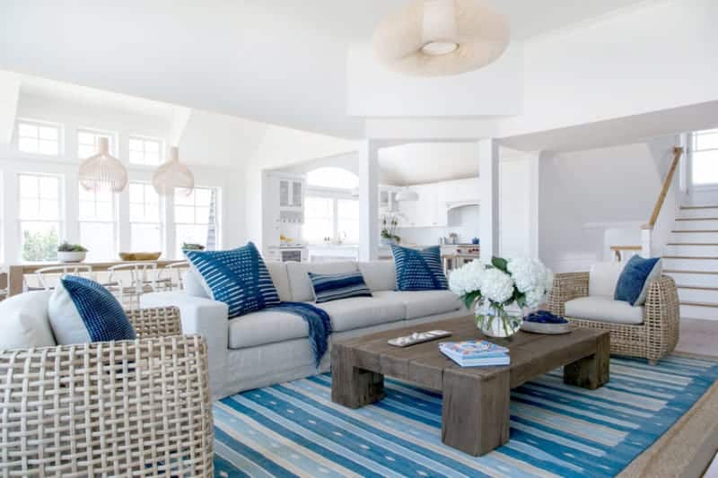 Beach style living room with a striped area rug and a rustic coffee table surrounded by gray seats that are accented with blue pillows. It is illuminated by a round chandelier along with natural light from the white framed windows.