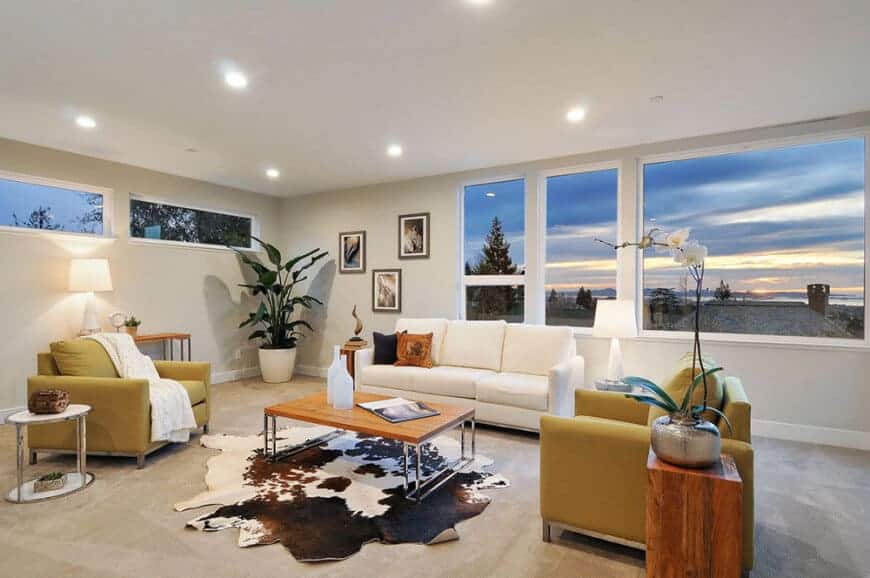 White living room with a sectional sofa and yellow armchairs surrounding a wooden coffee table on a cowhide rug illuminated by recessed ceiling lights.
