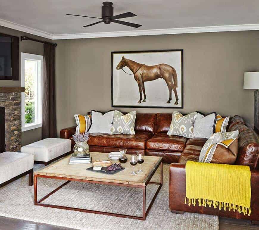 A horse painting hangs above the brown leather sofa filled with gray pillows and a yellow throw blanket. It is accompanied by a wooden coffee table and cushioned sits across the fireplace.