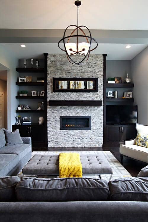 A gorgeous chandelier hangs over the tufted ottoman surrounded with cozy seats and a fireplace fixed on the brick pillar. It is accompanied by built-in shelvings filled with frames and decors.