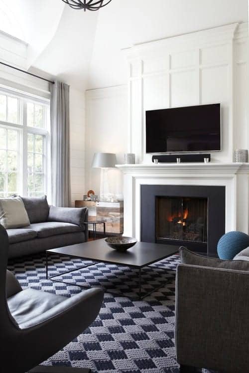 A checkered area rug adds a striking contrast to the white walls mounted with a flat-screen TV. This room features sleek seats and a metal coffee table that faces the fireplace.