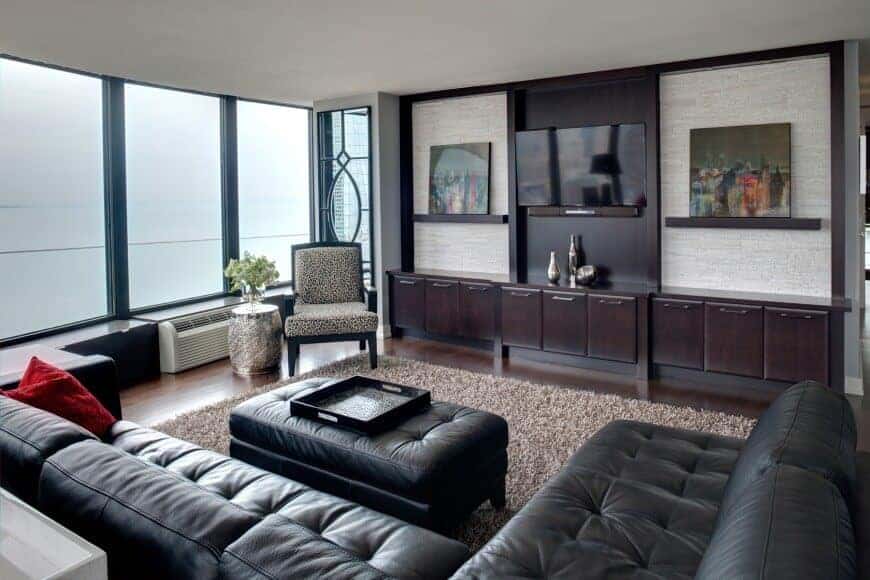 This living room offers a patterned armchair and black leather sofa paired with a matching tufted ottoman. It includes a shaggy rug and flat-screen TV mounted above the floating shelf.
